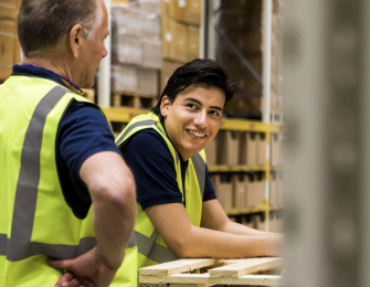 Page Image Smiling Workers in Hi-Vis Vests one leans on pallets and looks at the other who is facing away with hands on hips