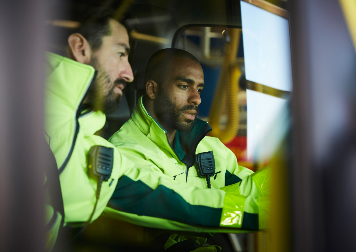 Page Image Men in Vehicle Cab with Hi Vis Jackets and Microphones attached to them, both men have a hand outstretched operating something out of frame