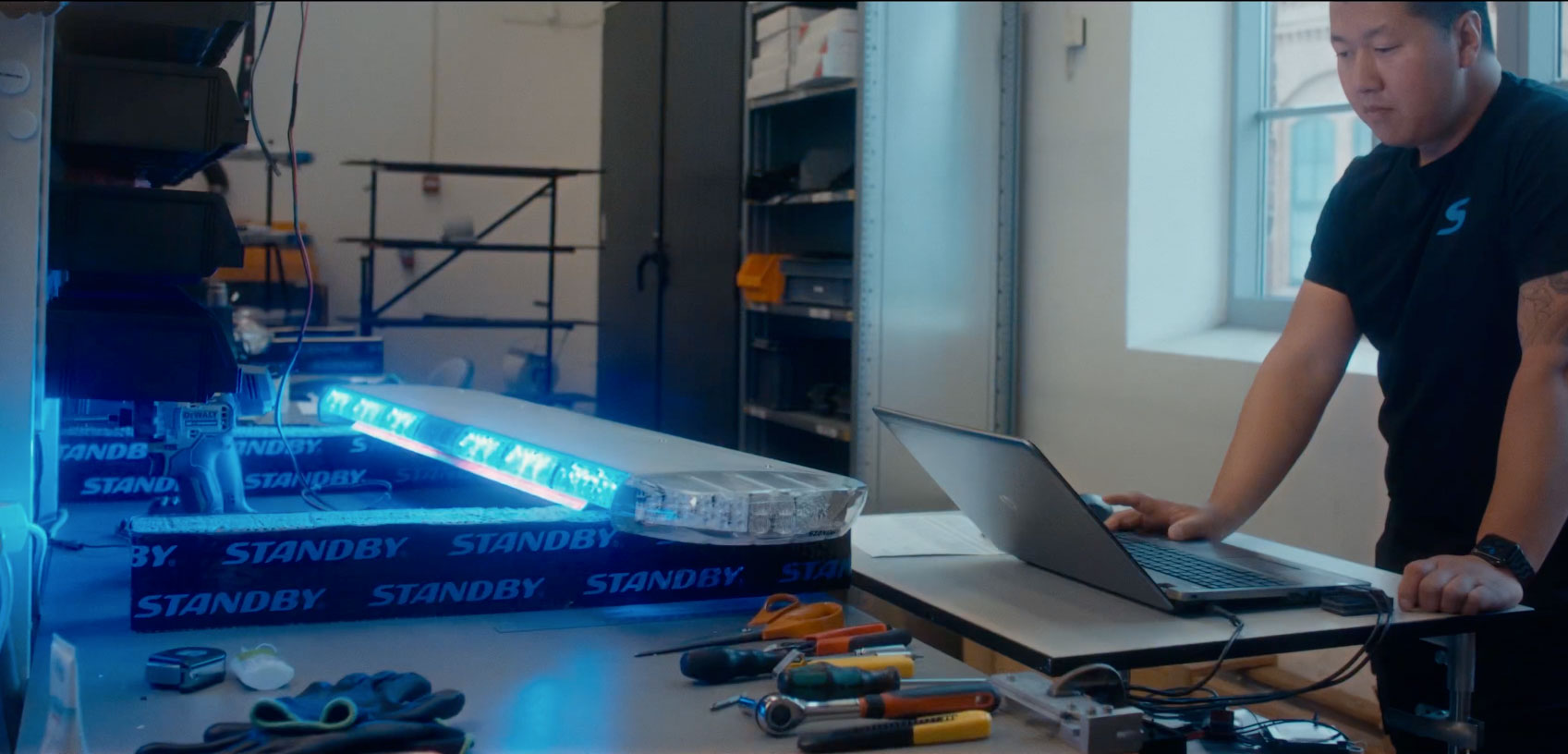 Hero Image Lightbar Engineer operates a laptop next to the W3 lightbar which is lit blue. Various tools are lined up on the bench in front of them