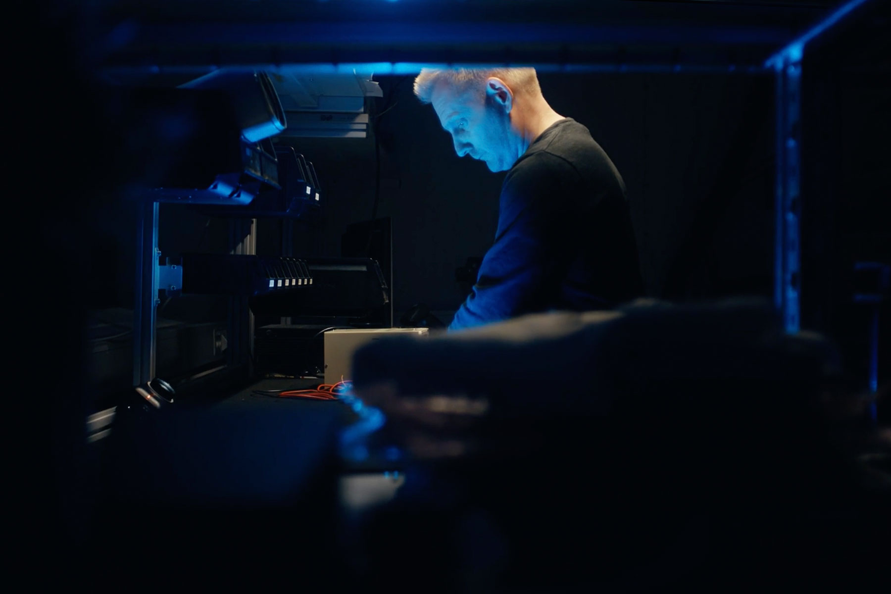 Page Image Engineer stands in dark room, lit up by the light they're working on, out of focus objects are dark in the foreground