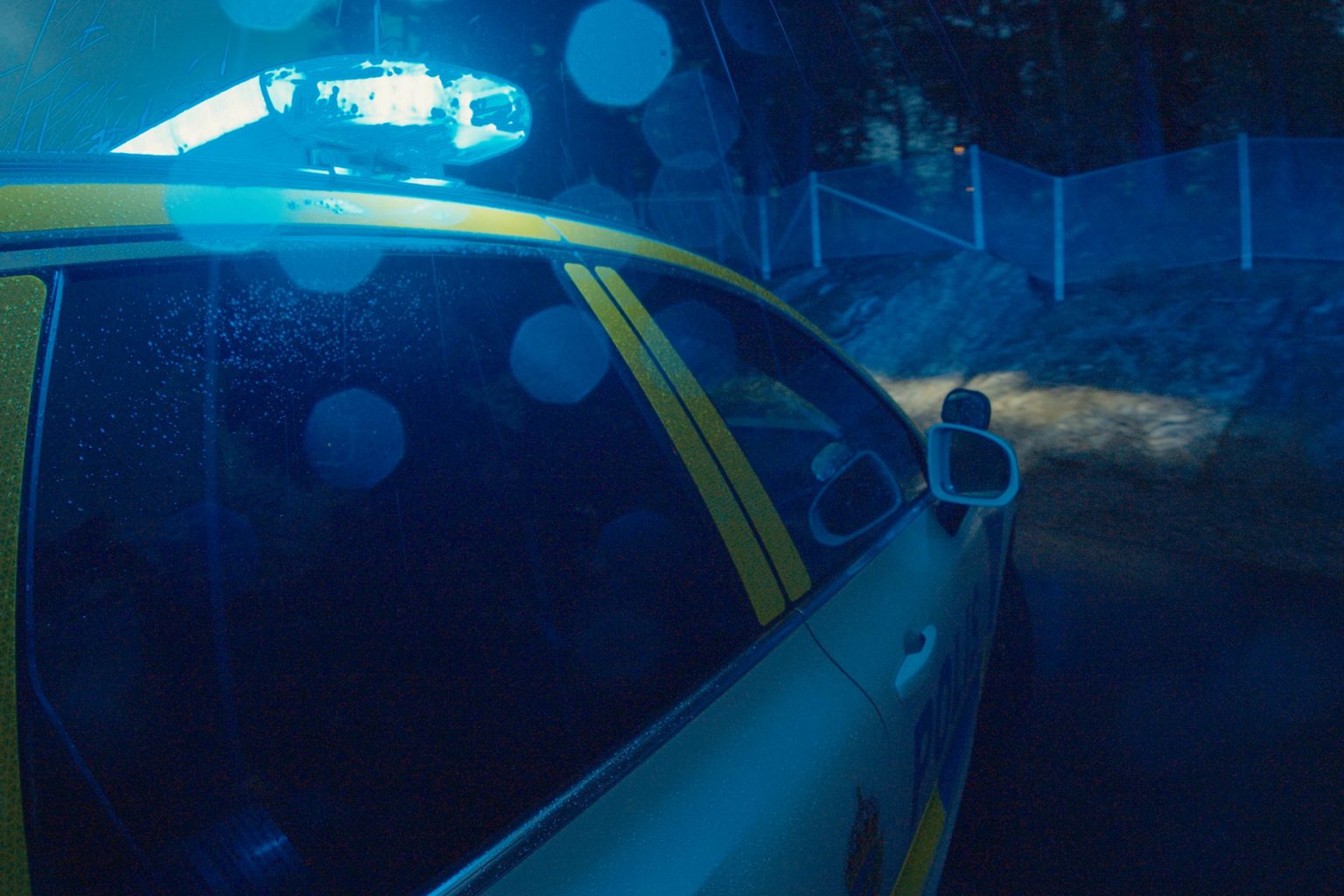 Page Image Lightbar on Car at Night Lit Blue, Screen Glares around lightbar on top of a white vehicle with yellow graphics, wooded location behind fence in background
