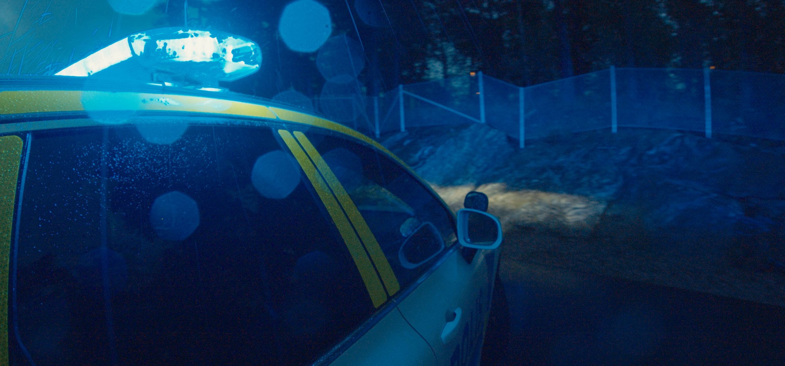 Hero Image Lightbar on Car at Night Lit Blue, Screen Glares around lightbar on top of a white vehicle with yellow graphics, wooded location behind fence in background