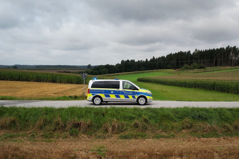 Page Image Police Van in Countryside Drives Along Paved Road Between Fields with Back Lightbar Lit Blue