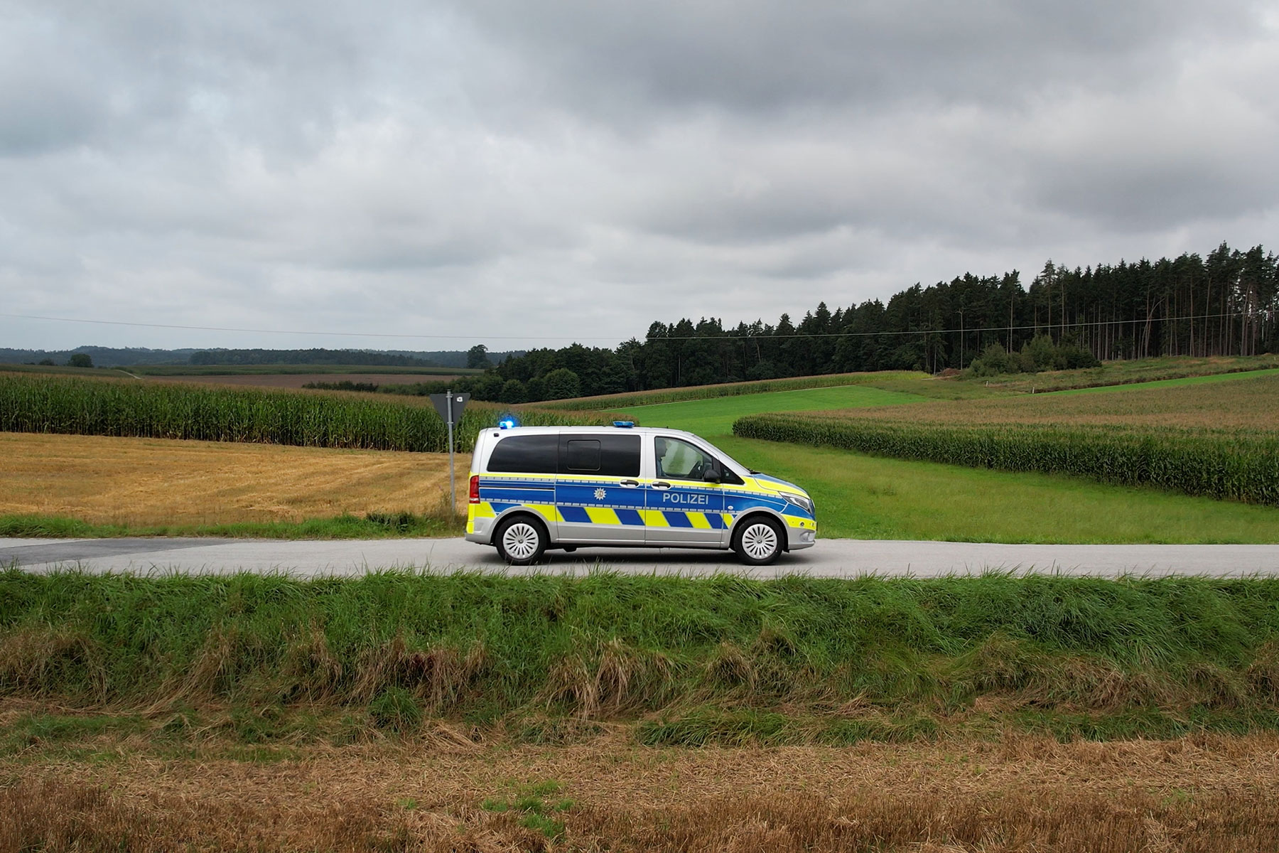 Page Image Police Van in Countryside Drives Along Paved Road Between Fields with Back Lightbar Lit Blue