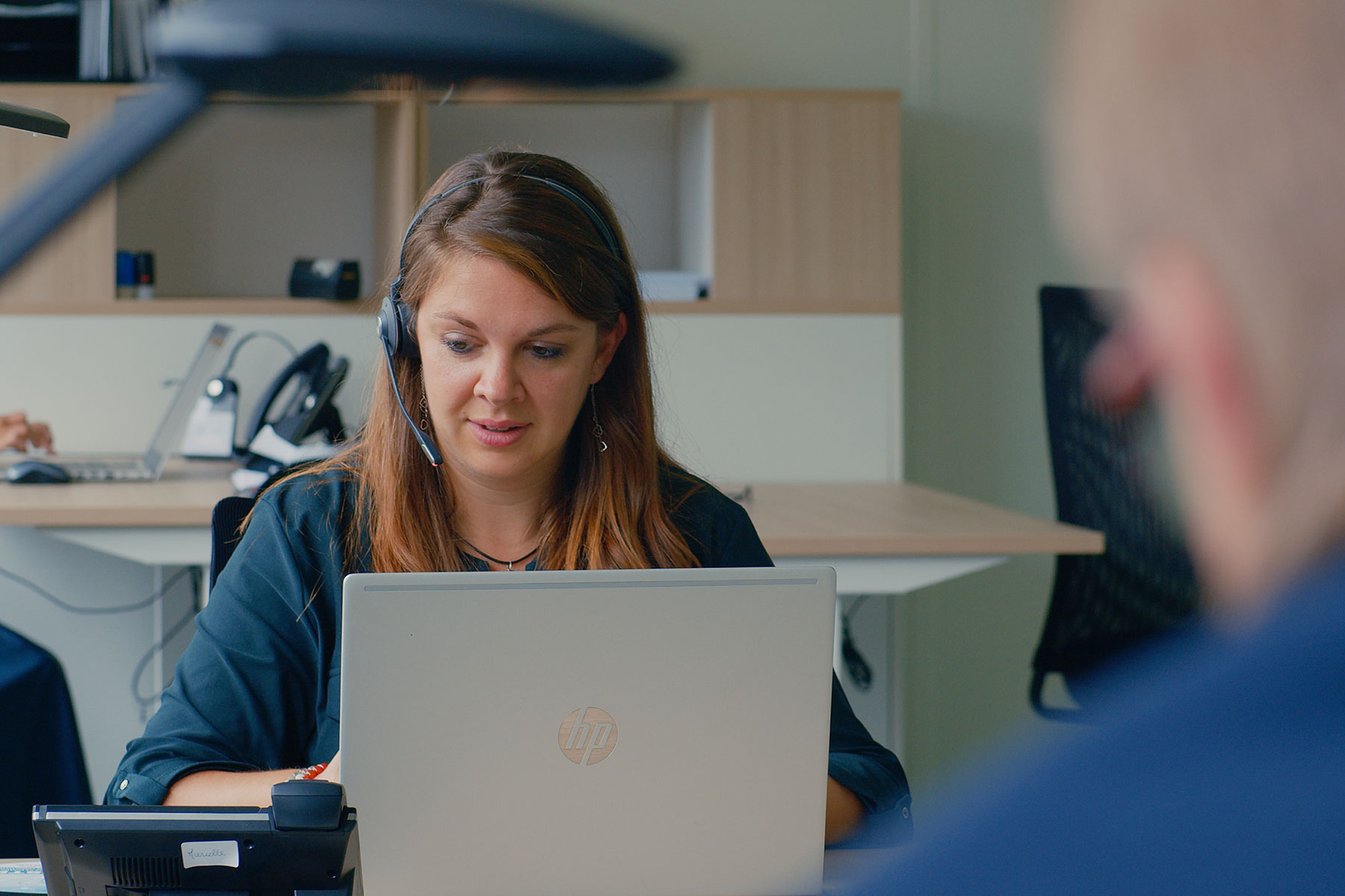 Page Image Person wearing headset is taking a phonecall while looking down at a laptop screen, over the shoulder shot of an out of focus figure in foreground