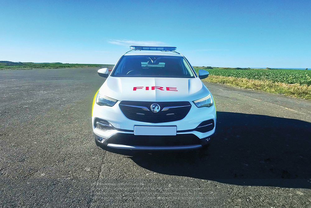Front view of white fire response vehicle with partially lit lightbar on cemented ground in front of flat scenic greenery with sea and sky in background