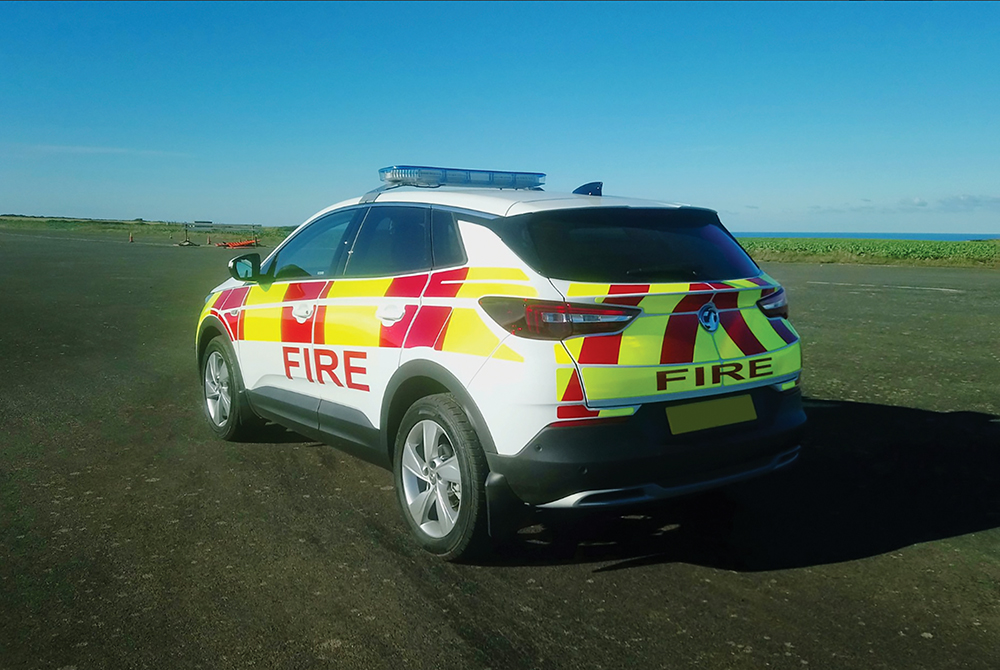 Angled reverse view of white fire response vehicle with partially lit lightbar on cemented ground in front of flat scenic greenery with sea and sky in background