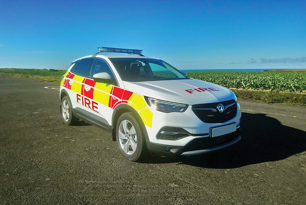 Angled front view of white fire response vehicle with partially lit lightbar on cemented ground in front of flat scenic greenery with sea and sky in background