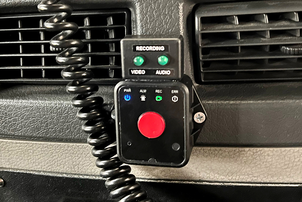 Red button on black square housing with two green unlit recording lights for video and audio with various indicators across top in front of vehicle vents with curly cable