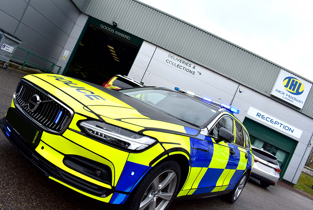 Angled view of police car with livery against front of building with Jack Hodson Ltd., Goods Inwards, Deliveries & Collections and Reception sign and silver vehicle