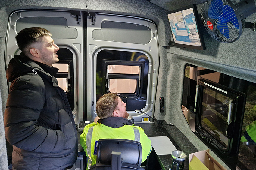 Safety Camera Van Interior Shot with two People Looking up Towards Monitor in Upper Lefthand Corner of the Photo, One Person Seated, One Standing Behind Them