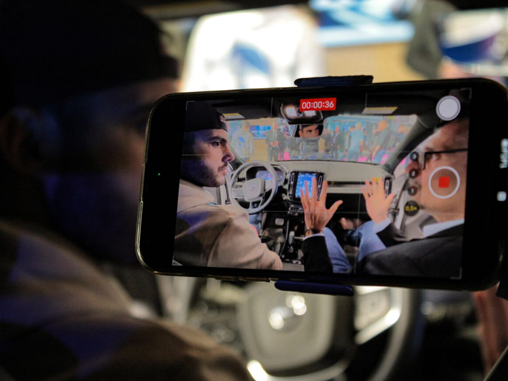 Internal shot focused on a mobile phone screen showing two people in the front of the car talking and being recorded.