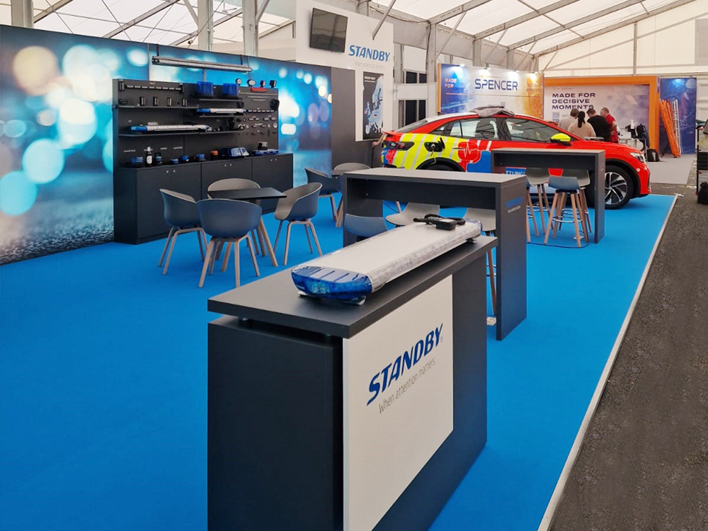 Standby Group exhibition stand with various products and seated areas, red response vehicle in background, lightbar covered.