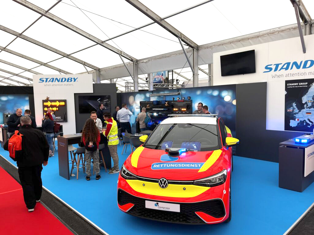 Image of Standby exhibition booth at RETTmobil International 2024 indoor trade show, taken at an abgle view. Red emergency vehicle runs perpendicularly to the camera angle, showing the front of the vehicle. Groups of people stand around in discussion.