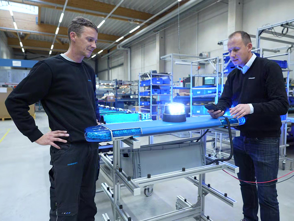 Two male workers stand either side of a blue unlit lightbar with a blue beacon lit up on top, looking down at the product.