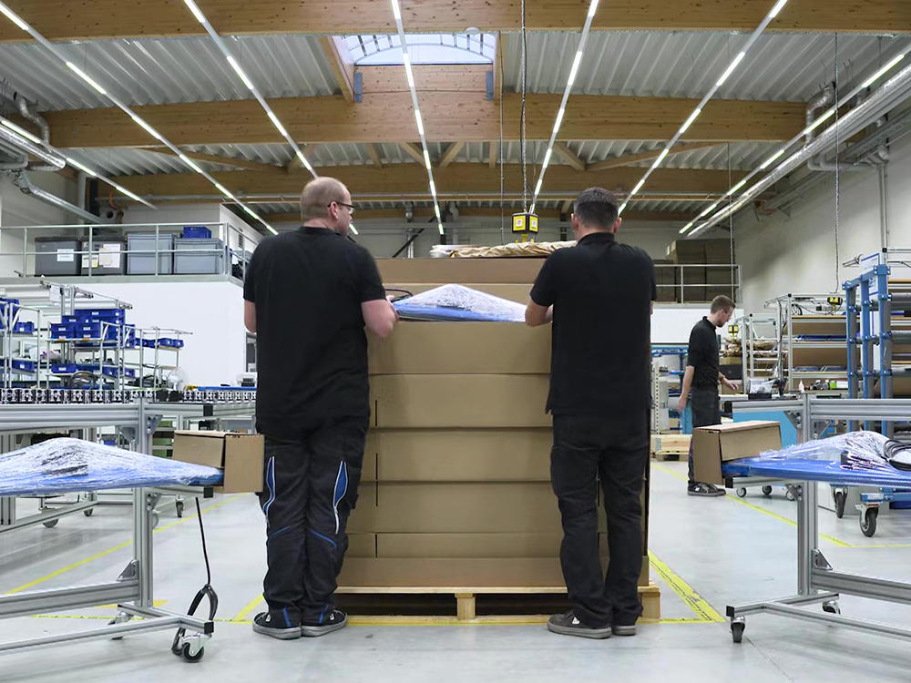 Two male workers stand with their backs to the camera, lowering a wrapped product into a cardboard box in a bright, neat and clean factory environment.