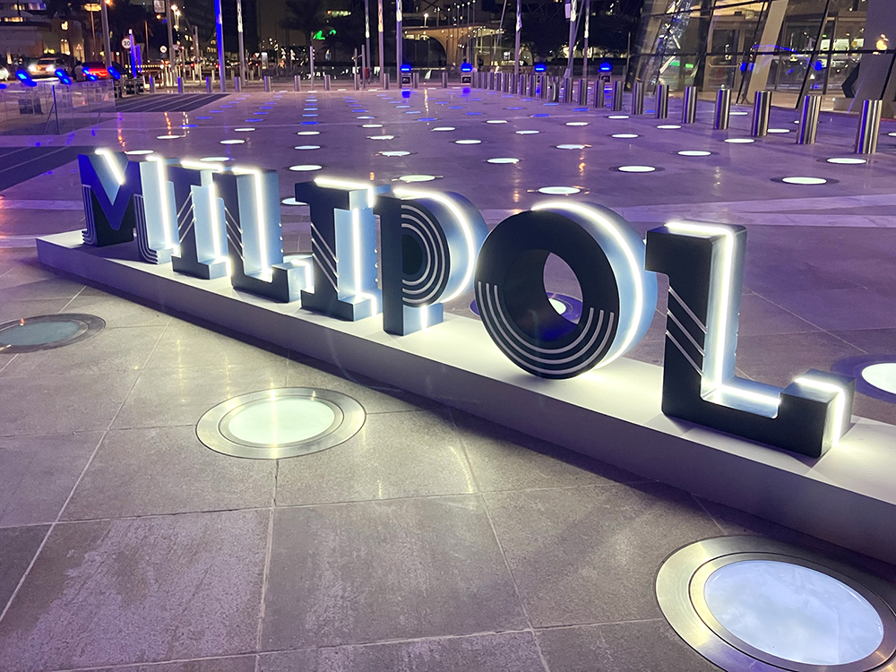 Angle view of a 3d lit up Milipol sign mounted on the pavement in an external square outside of the DECC Doha, taken at night.