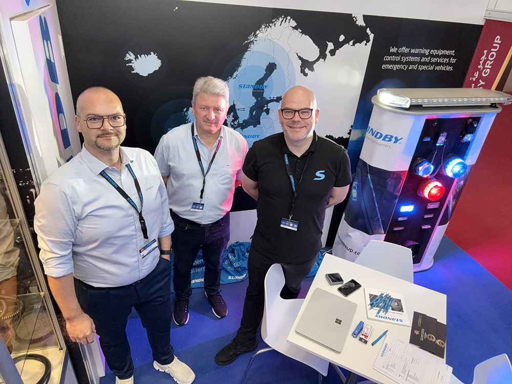 Three smart male employees on the Standby exhibition stand, taken at a high angle at Milipol Qatar 2024. They are smiling and looking at the camera.