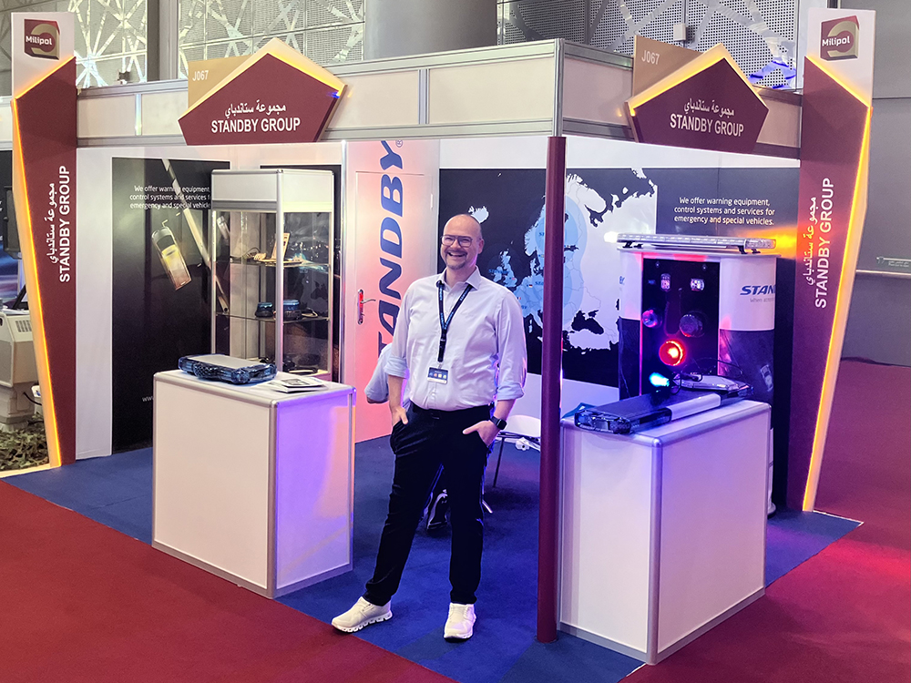 A smart male employee stands on the Standby Group exhibition stand with his hands in his pockets at Milipol Qatar 2024. The whole stand is in view, taken at an angle.