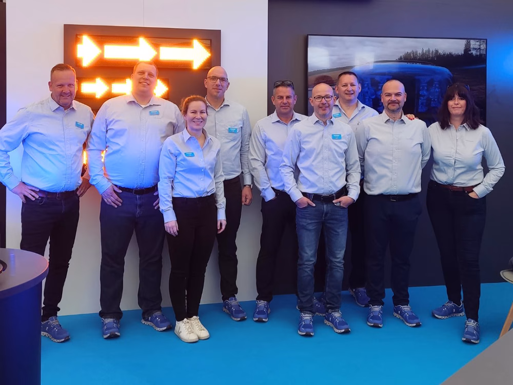 Standby Germany team on the RETTmobil Standby stand all wearing light blue shirts with dark jeans and blue trainers. They stand in front of a message display board and TV screen.