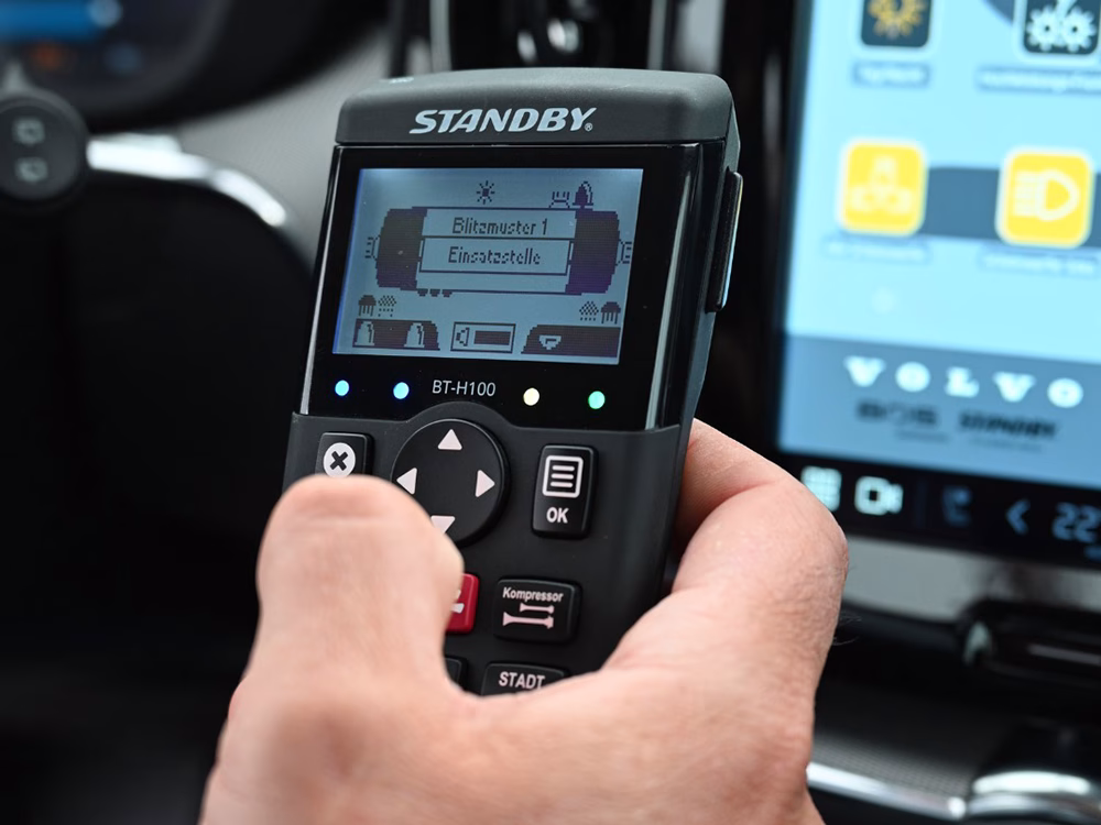 Standby BT-H100 Contol Unit held in hand in front of an out of focus Volvo infotainment screen.