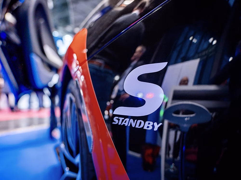 Closely cropped view of the Standby and Standby 'S' logos on a black McLaren next to the wheel arch. Reflections of the Standby Group stand and visitors at Milipol Paris 2025.