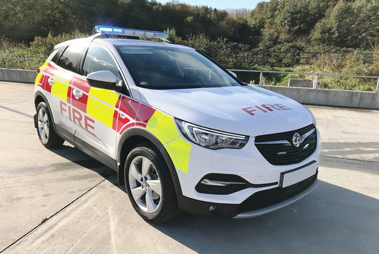 Angle view of white fire response vehicle with half lit lightbar on cemented ground in front of scenic greenery