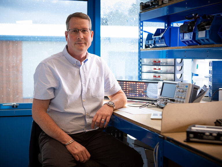 Smartly dressed worker is looking at camera and smiling, sitting at a workbench with various electronic products in front of a window