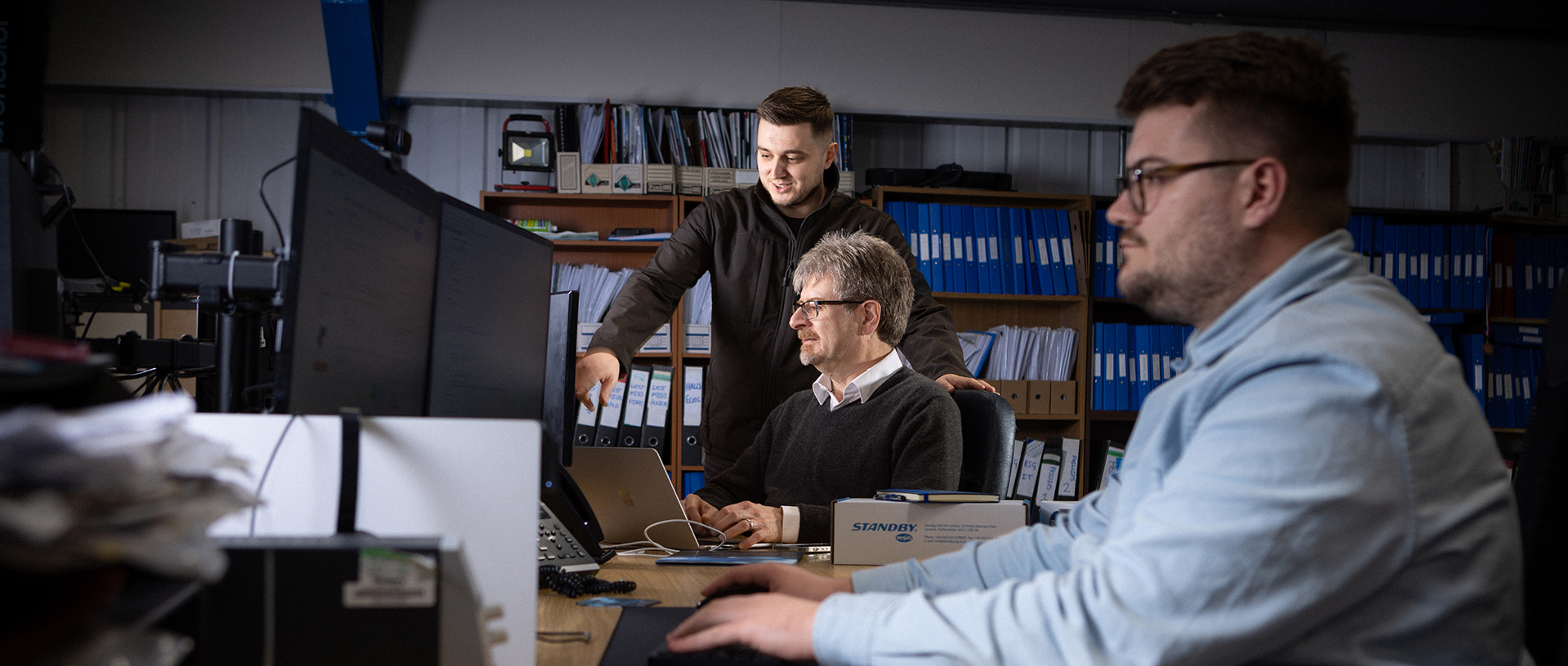 Two smartly dressed workers are in focus in the background against a wall of various folders, one is seated at their desk and the other is standing behind them pointing at their PC screen, a third worker is out of focus sitting side on in the foreground working at their PC