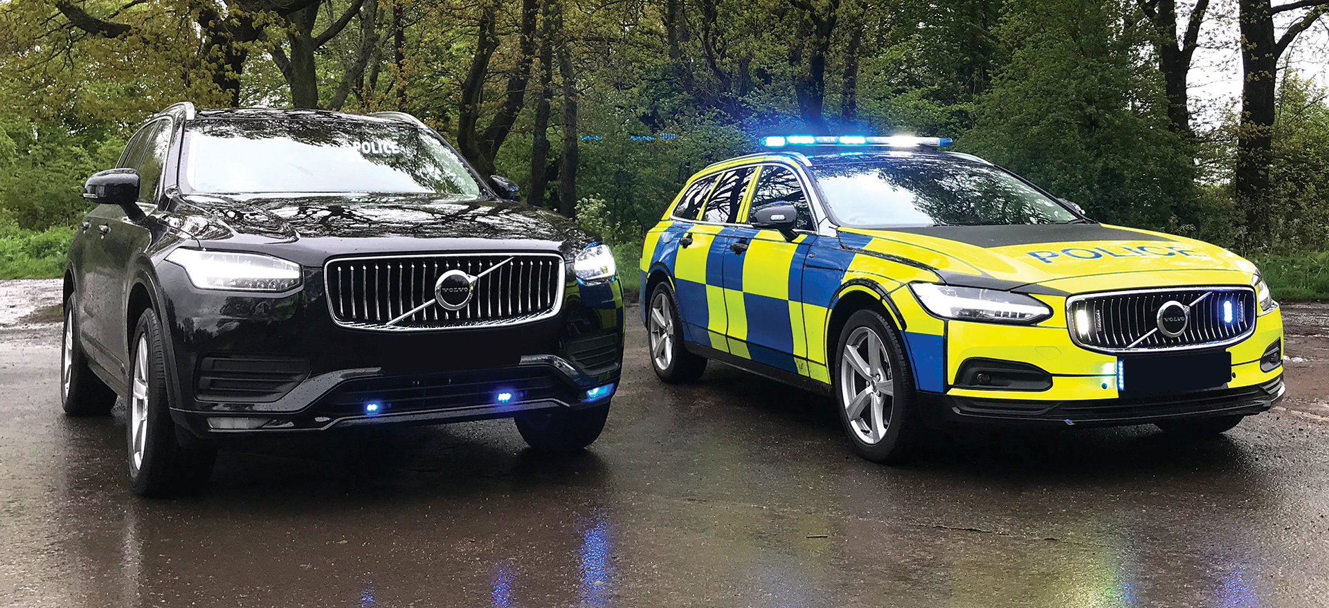 Black unmarked Volvo parked next to marked Police Volvo with livery, both at an angle, against wooded background, emergency lights reflecting off wet tarmac floor