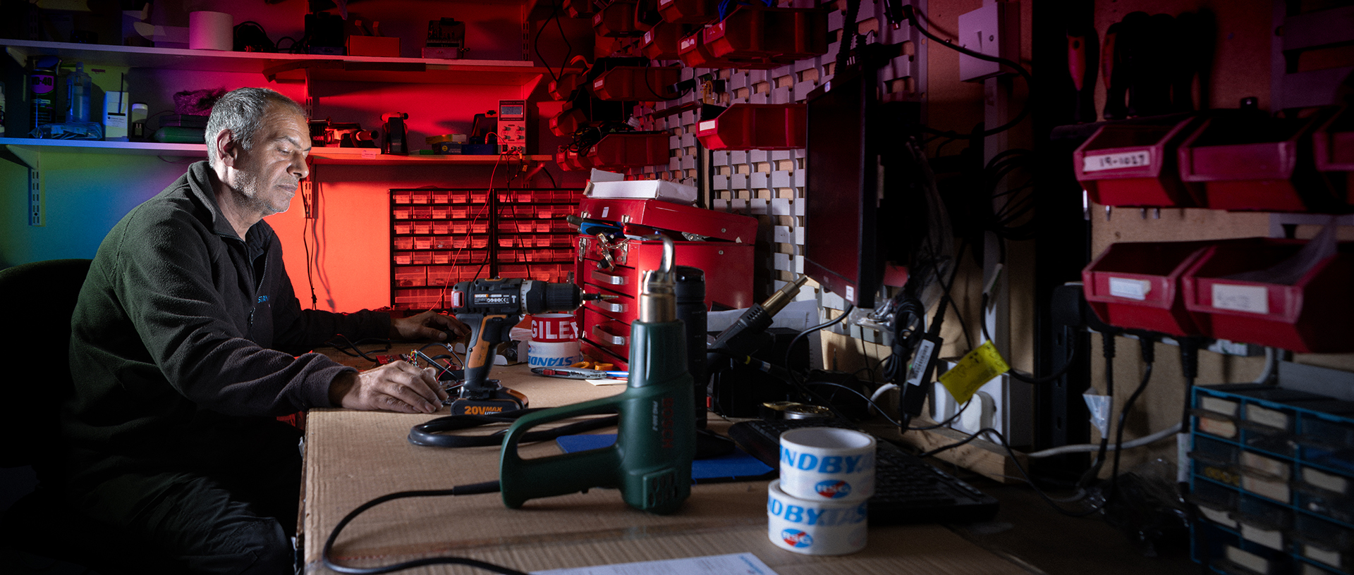 Side on of a worker seated at workbench against a red lit background working on a technical task in a workshop