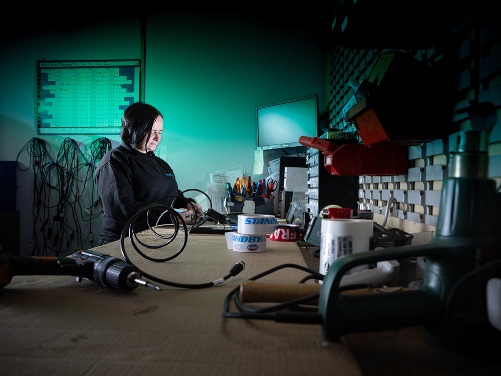 Worker is completing a technical task at their workbench in a workshop environment, looking down at what they're doing, a cable is coiled in the foreground reaching towards the camera, the back wall is lit green