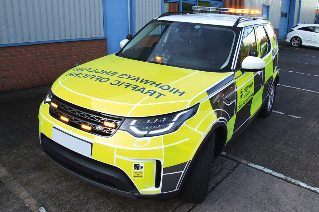 Angle View of Highways England Traffic Officer Vehicle with Amber Grille Lights, Lightbar and Dash Light on Car Park in Front of Industrial Unit Building