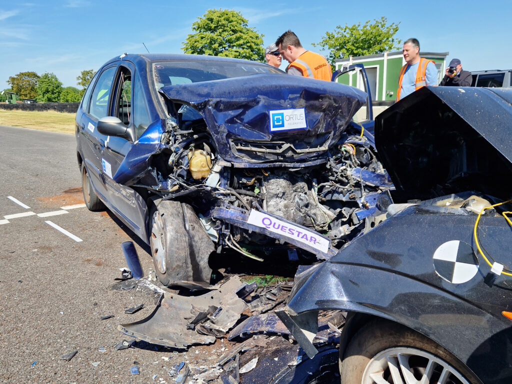 Close up of two cars subjected to controlled collision, bonnets of both are crumples with debris scattered around and hanging off. People in orange hi-vis jackets are in the background of the image as well as a photographer.