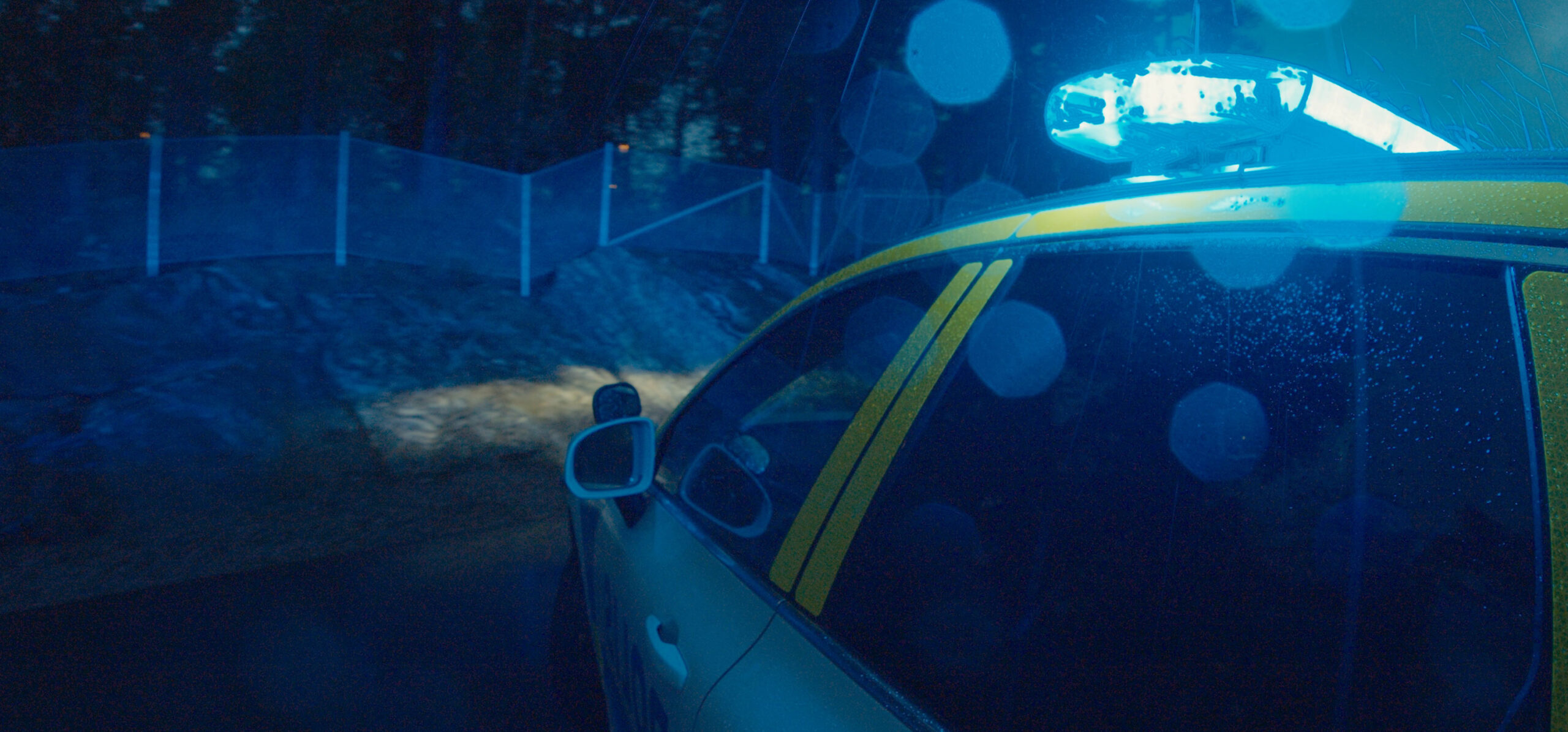 Lightbar on Car at Night Lit Blue, Screen Glares around lightbar on top of a white vehicle with yellow graphics, wooded location behind fence in background
