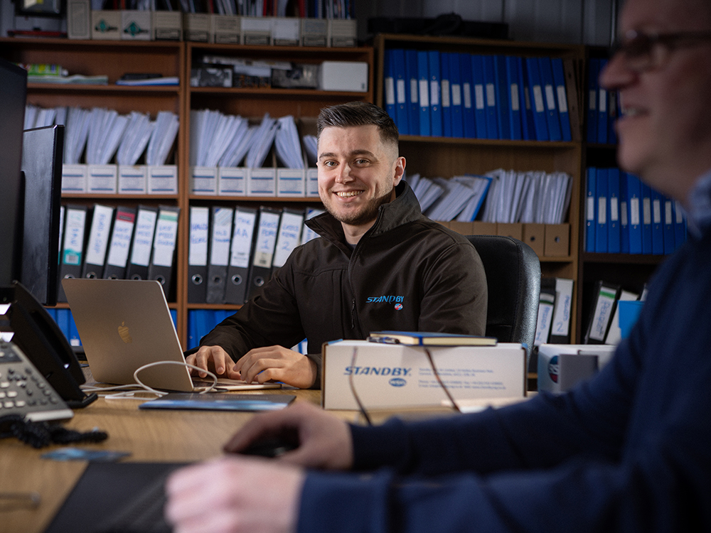Worker in black Standby UK fleece sits at a desk, looking toward the camera and smiling. Their hands are poised at a laptop keyboard, another worker sits side on in the foreground out of focus, in the background is a bookcase filling the frame with files and folders