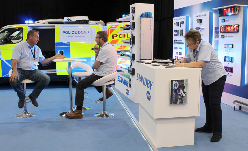Side view of staff on Standby's NAPFM stand, two sit in conversation at a high desk and one leans on centre table writing
