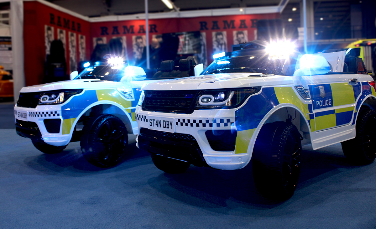 Standby Demo Cars - 2 x children's ride-on RC cars with police livery, blue and white lights and 'ST4NDBY' licence plates, RAMAR stand is out of focus in the background