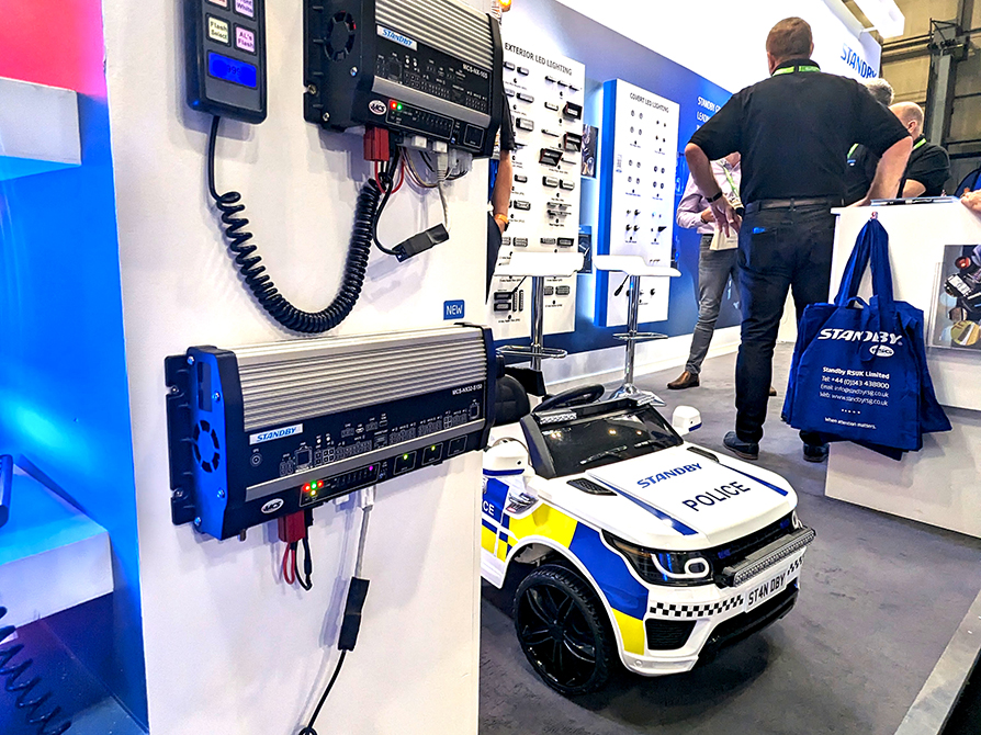 Close up of Standby RSG stand at The Emergency Services Show 2023 showing new NX range in the left foreground, a children's ride on Police car and Standby RSG branded tote bags in the centre midground and a crowd of people around lighting boards in the background