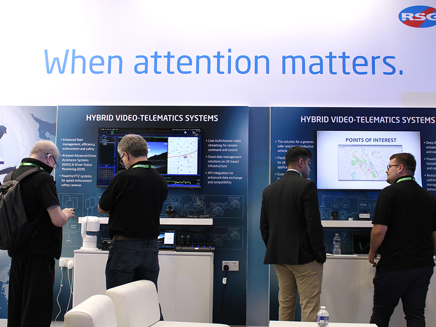 Close up of the Standby RSG stand at The Emergency Services Show 2023 with 4 people in front of 2 Hybrid Video-Telematics Systems boards with their backs to the camera. Large text on the top of the stand reads 'When attention matters.'