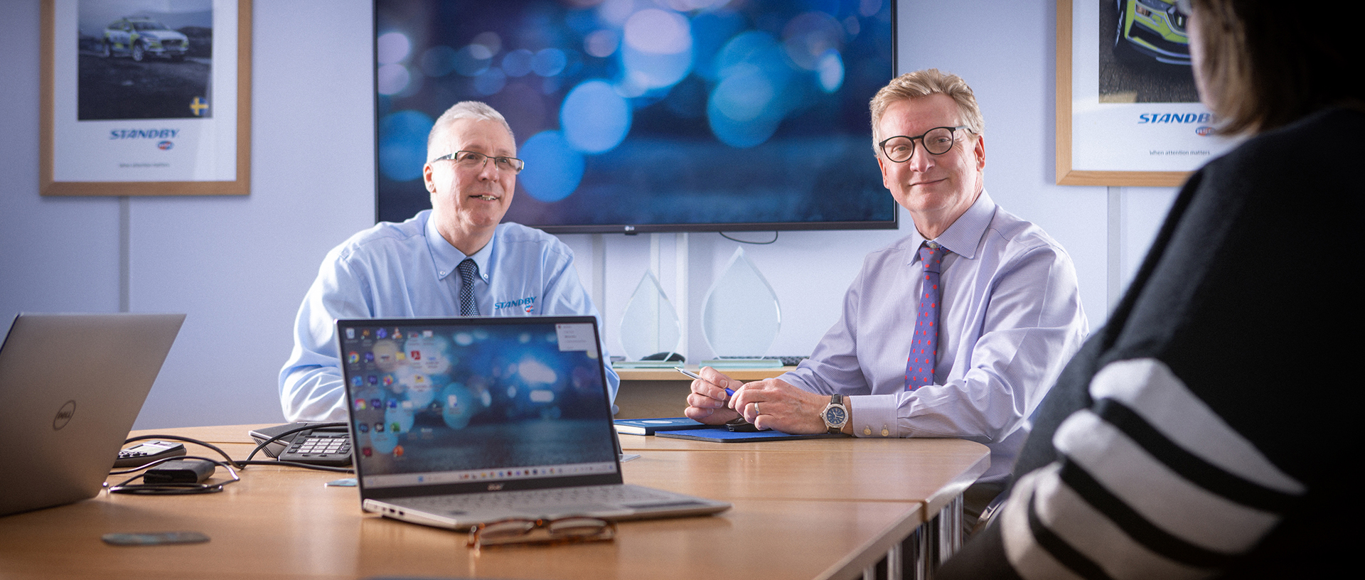 Two smartly dressed workers sit behind table looking to someone off shot to the righthand side, image is taken over their shoulder, the setting is a boardroom, a laptop is open and on, facing the camera in the middle of the image