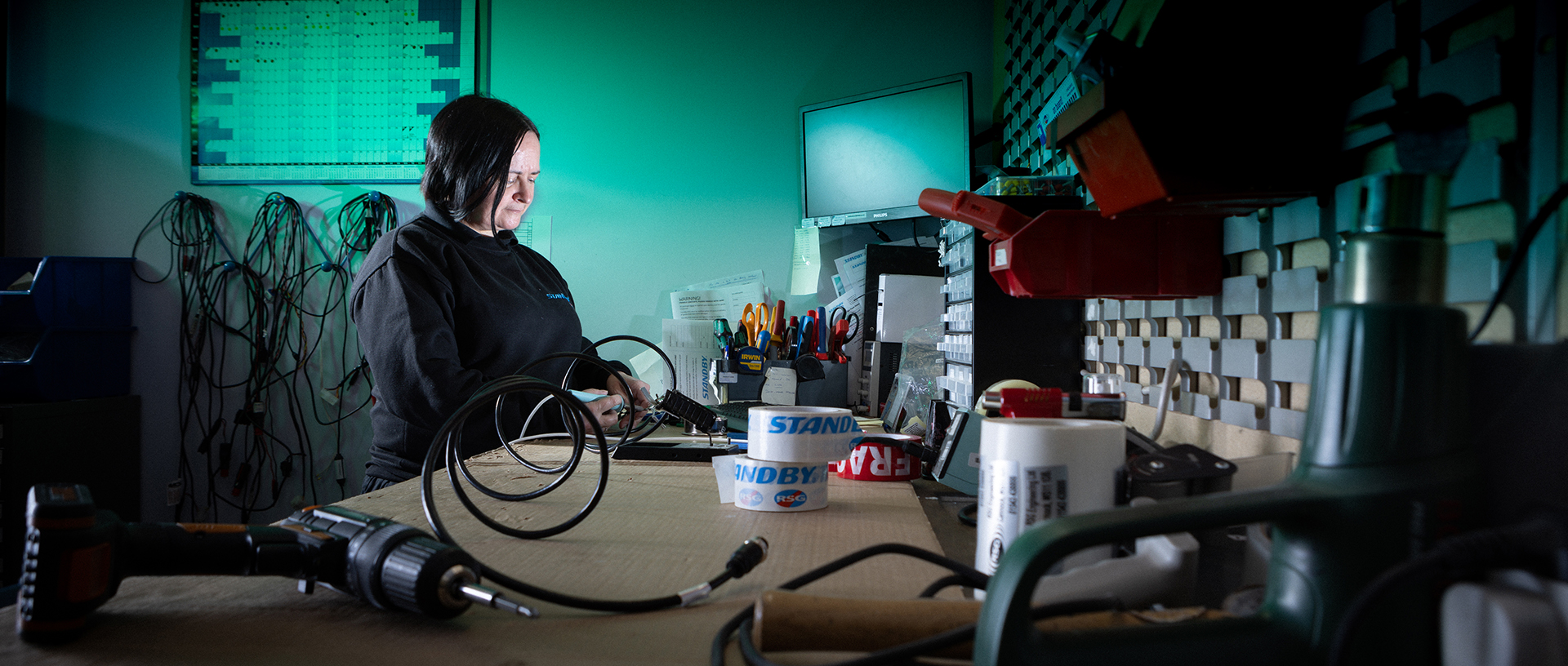Worker is standing at workbench soldering and looking down at her work, the workbench runs towards the camera with various tools and equipment on. The background of the image is lit green.
