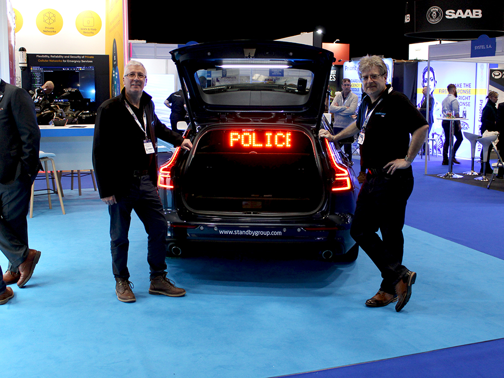 Two Standby RSG colleagues stand either side of an open car boot with an LED Matrix sign reading 'Police' in amber lights on Standby RSG exhibtion stand.