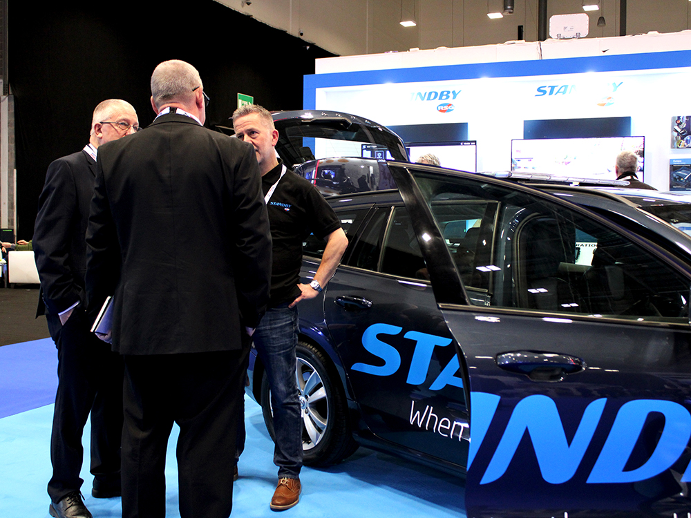 Candid shot of Standby colleague speaking with two men in suits next to the demonstration car on the Standby RSG exhibition stand. The front door and car boot are open.