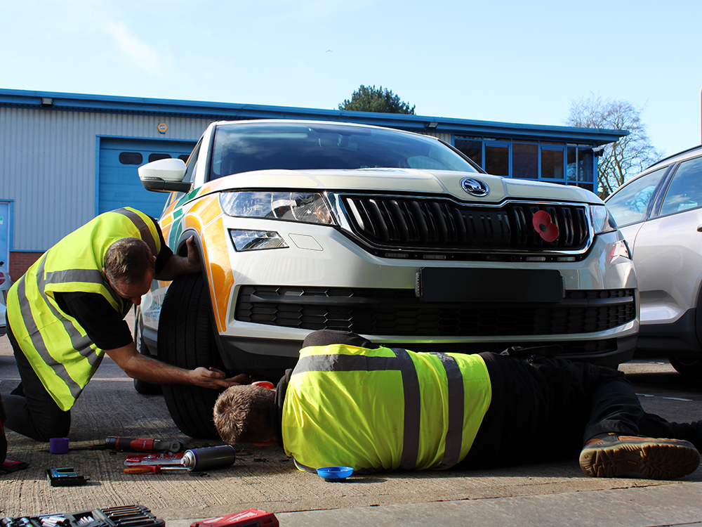 Two workers in hi-vis vest work on the floor on the front end of a response car, one is kneeling the other lying down with their back to us, accessing the underside of the vehicle.