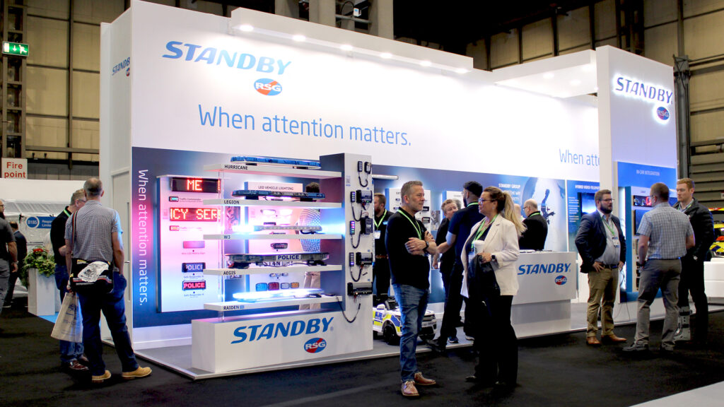 Angle shot of the Standby RSG NAPFM stand at The Emergency Service Show 2023. The stand is lit up and has lots of staff and visitors chatting.