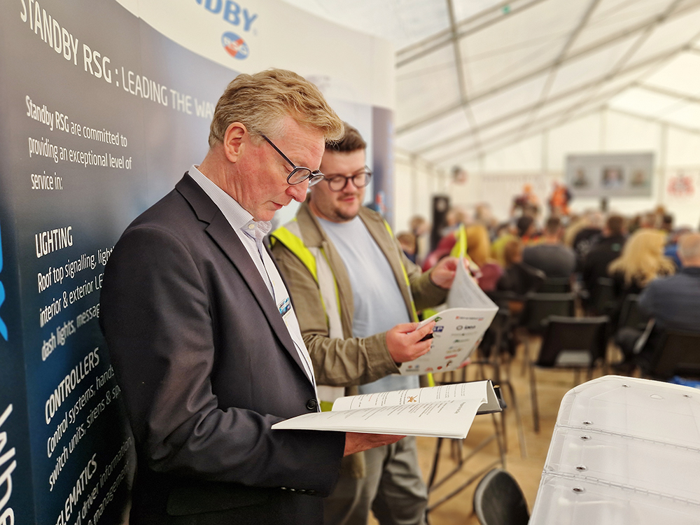 A smartly dressed business man stands next to an employee in a hi-vis vest on a small exhibition stand in a white marquee. Both look down at event brochure.