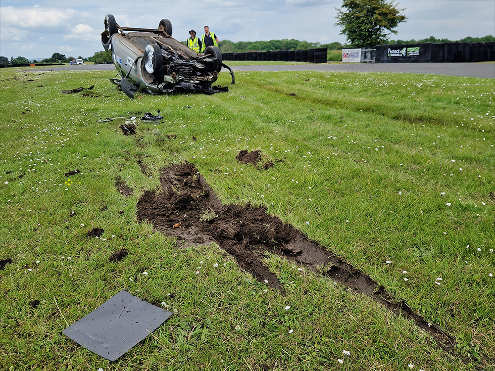 Damaged overturned silver vehicle in the distance, shot focuses on the grass with a gouge missing from it in the direction of the vehicle. Two men in hi-vis vests stand near the vehicle.