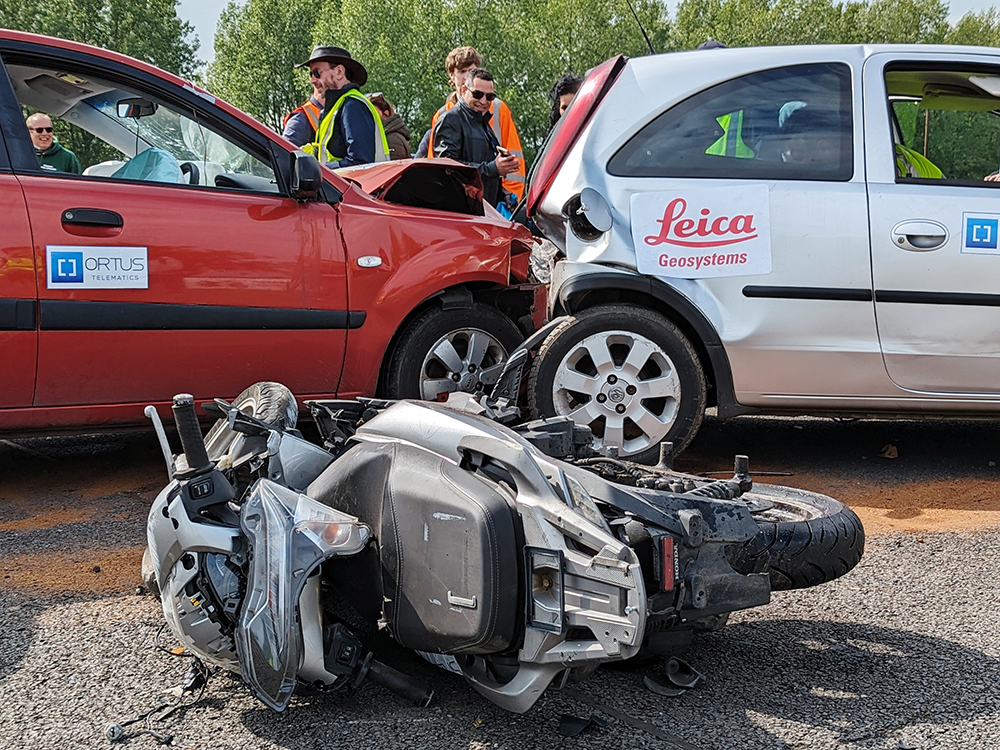 Closely cropped photo of a red vehicle which has crashed into the back of a silver car, both heavily damaged. A motorbike is overturned on the ground in front of the crash and a group of people are gathered in the distance.