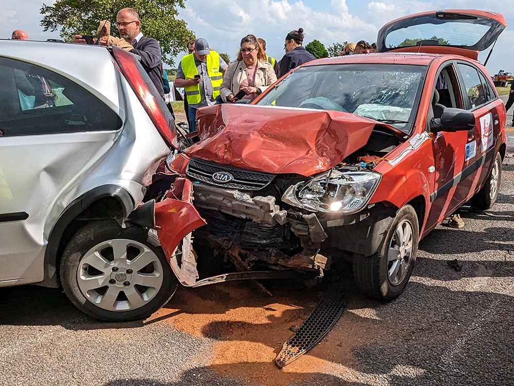 A red Kia with a damaged bonnet and engine bay has crashed into the back of a silver vehicle. The headlight is on. A group of people gather in the background, looking at the crash. Sand has been scattered on the tarmac under the vehicles.